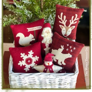 Peut inclure: Un panier en osier blanc rempli de coussins en feutre rouge décorés de motifs de Noël brodés. Les coussins présentent un flocon de neige, un renne, un écureuil, un bonhomme de neige, un ours polaire et le Père Noël. Le panier est posé sur une surface en bois avec un sapin de Noël vert en arrière-plan.