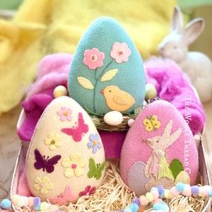 May include: Three felt Easter eggs in a pink floral suitcase. The eggs are decorated with embroidered flowers, butterflies, a bird, and a bunny. The suitcase is filled with straw and has a handle.