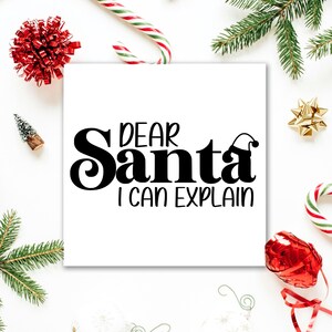 May include: A white card with black text that reads "Dear Santa I can explain". The card is surrounded by Christmas decorations, including candy canes, ornaments, and a small Christmas tree.