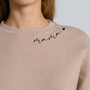 Puede incluir: Una sudadera de color marrón claro con la palabra "mama" y un pequeño corazón impreso en el hombro izquierdo.