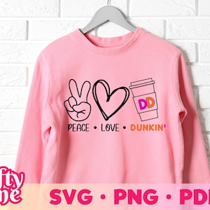 Könnte beinhalten: Rosafarbener Pullover mit einem Peace-Zeichen, einem Herzen und einem Dunkin' Donuts-Kaffeebecher. Der Text lautet "Peace. Love. Dunkin'."