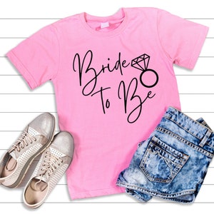 Puede incluir: Una camiseta rosa brillante de manga corta con el texto negro Bride to Be y un gráfico de un anillo de diamantes. La camiseta se muestra junto a un par de zapatillas plateadas perforadas y unos pantalones cortos de mezclilla azul.
