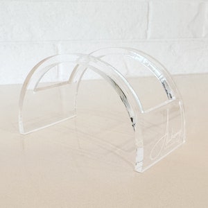 May include: A clear acrylic stand with a curved design. The stand has two slots for holding items, and the word "Alchemy" is etched on the side.