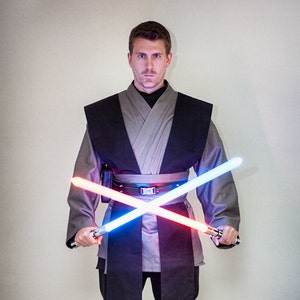 May include: A person dressed as a Jedi Knight, wearing a grey and black robe, holding two lightsabers, one blue and one red, crossed in front of them.