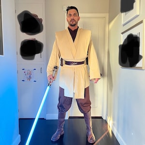 May include: A person dressed as a Jedi Knight, wearing a tan robe and brown pants, holds a blue lightsaber in their right hand. The person is standing in a hallway with a white door and brown wood floor.