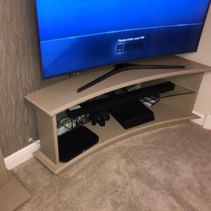 Puede incluir: Un centro de entretenimiento curvo de color beige con un estante de vidrio y dispositivos electr&oacute;nicos negros, que incluyen una barra de sonido, consolas de juegos y un televisor que muestra una pantalla azul con el texto "Por favor, ingrese su PIN".