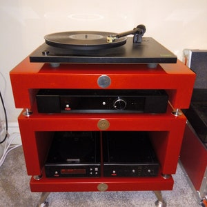 May include: A red wooden audio rack with three shelves. The top shelf has a black turntable with a record on it. The middle shelf has a black amplifier. The bottom shelf has two black CD players.