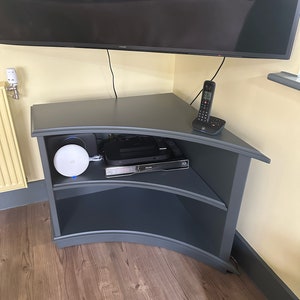 Puede incluir: Un soporte de TV de esquina gris oscuro con dos estantes. El soporte tiene un frente curvo y est&aacute; hecho de madera. Hay un router blanco, una caja de cable negra y un tel&eacute;fono negro en el estante superior.