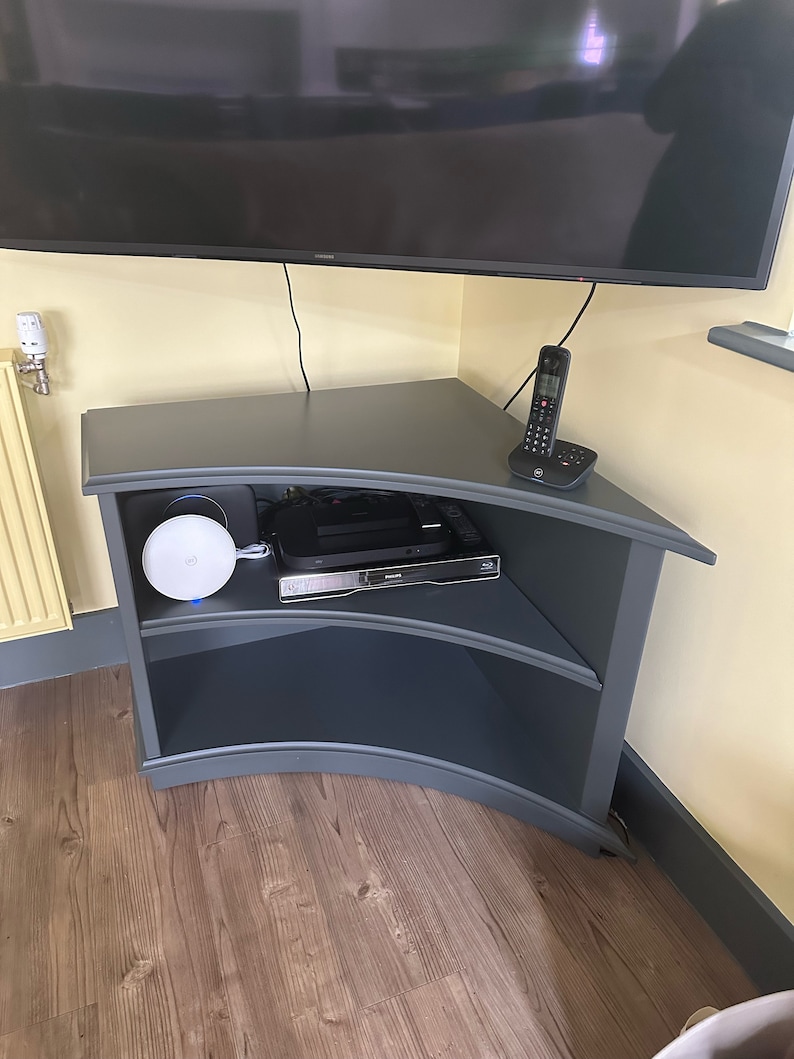 Puede incluir: Un soporte de TV de esquina gris oscuro con dos estantes. El soporte tiene un frente curvo y est&aacute; hecho de madera. Hay un router blanco, una caja de cable negra y un tel&eacute;fono negro en el estante superior.