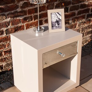 May include: A white bedside table with a mirrored drawer and a crystal lamp with a clear glass shade. The table has a small shelf below the drawer.