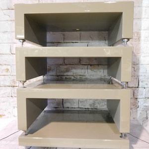 May include: A three-tiered audio equipment stand made of light grey wood with a glossy finish. The stand has a unique design with open sides and rounded edges. Each shelf is supported by four small metal feet.