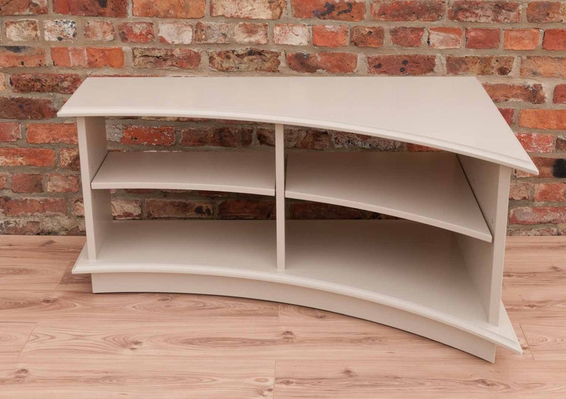Puede incluir: Un centro de entretenimiento curvo de color gris claro con tres estantes. La unidad est&aacute; hecha de madera y tiene un dise&ntilde;o moderno.