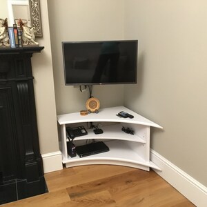 Puede incluir: Un soporte de TV de esquina blanco con dos estantes. El soporte tiene un frente curvo y est&aacute; hecho de madera. Un televisor de pantalla plana est&aacute; montado en la pared sobre el soporte.