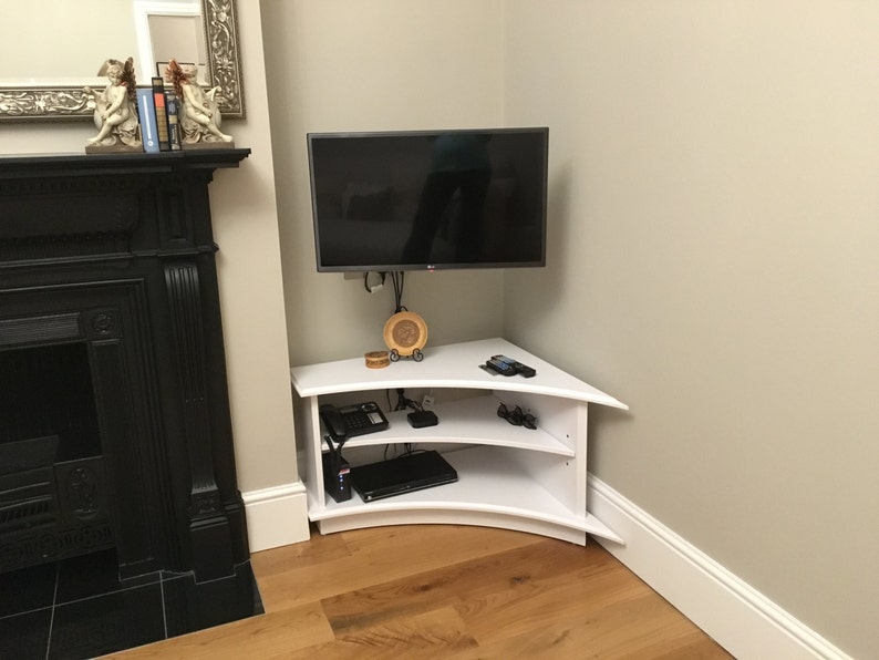 Puede incluir: Un soporte de TV de esquina blanco con dos estantes. El soporte tiene un frente curvo y est&aacute; hecho de madera. Un televisor de pantalla plana est&aacute; montado en la pared sobre el soporte.