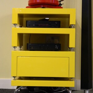 May include: A yellow entertainment unit with a turntable, a CD player, and a receiver on the shelves. The unit has a drawer and sits on four chrome legs.