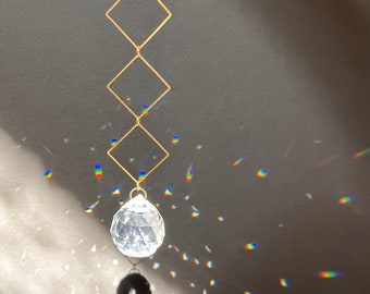 May include: A crystal ball hanging from a gold chain with three square frames. The crystal ball is casting rainbows on a gray surface.