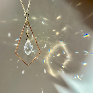 May include: A gold necklace with a diamond-shaped pendant featuring a clear crystal drop. The necklace is photographed against a white background with light reflecting off the crystal.