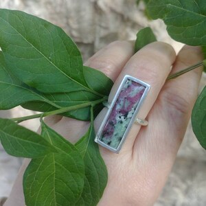 Op de afbeelding: Een rechthoekige ring met een zetting van gepolijst zilver die een platte, langwerpige steen bevat. De steen vertoont een gevlekt patroon van diep magenta, bosgroen en zwarte spikkels, wat een duidelijk contrast vormt met het metaal.