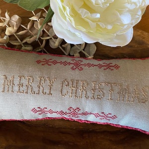 Könnte beinhalten: Ein beigefarbenes Kissen mit rosa Rand und den Worten "Merry Christmas", die in braunem Garn gestickt sind.