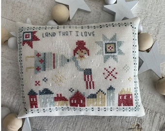 Puede incluir: Una almohada blanca con un diseño de punto de cruz que presenta una figura roja, blanca y azul con el texto "Land That I Love".