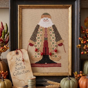 Puede incluir: Un bordado en punto de cruz enmarcado de una figura de Santa con un abrigo estampado en tonos verde, beige y rojo. Una etiqueta de papel beige con el texto AC Sep Santa Collection está unida al marco, junto a un carrete de hilo y tijeras.