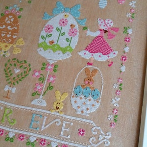 May include: Cross stitch pattern featuring a pastel colored Easter scene with a girl holding a basket of eggs, a bunny, and a decorated egg. The text "REVE" is stitched below the scene. The text "Cuore e Batticuore" is stitched at the bottom of the pattern.