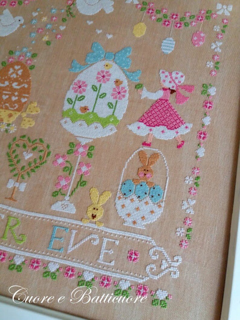 May include: Cross stitch pattern featuring a pastel colored Easter scene with a girl holding a basket of eggs, a bunny, and a decorated egg. The text "REVE" is stitched below the scene. The text "Cuore e Batticuore" is stitched at the bottom of the pattern.