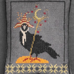 Pode incluir: Um corvo preto usando um chapéu de bruxa vermelho e amarelo está em uma plataforma amarela e laranja. O corvo está segurando um bastão com uma lua crescente no topo. O fundo é um tecido cinza com uma borda de estrelas amarelas.