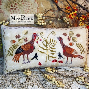 May include: Cross stitch pattern featuring two turkeys with red berries, green leaves, and brown acorns. The pattern is on a white pillow with a scalloped edge. The text "Miss Prim. Cross Stitch Pattern" is in the top left corner.