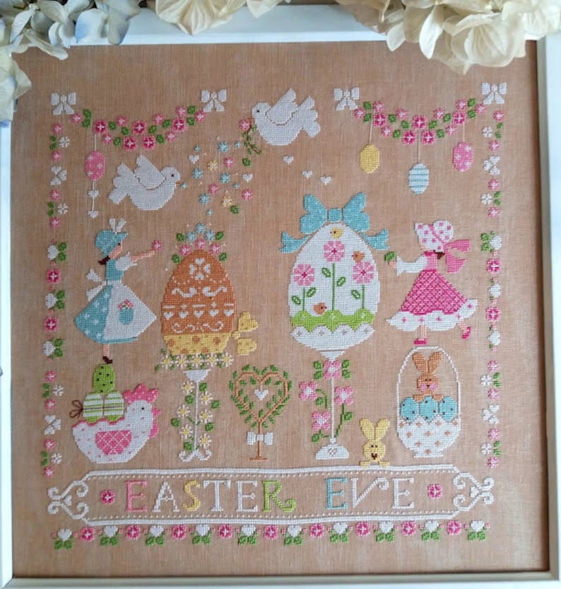 May include: A cross-stitch embroidery design featuring a pastel colored Easter scene with bunnies, birds, eggs, and flowers. The text "Easter Eve" is stitched in the bottom center of the design.