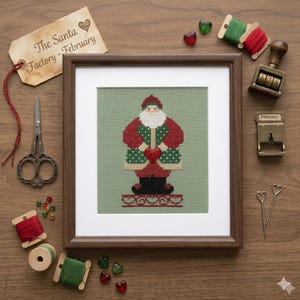 May include: A framed cross-stitch embroidery of Santa Claus holding a red heart, centered on sage green fabric. The design features red, green, white, and black thread. Surrounding items include vintage scissors, thread spools, and a tag labeled The Santa Factory - February.