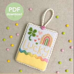 May include: A rectangular cross-stitch ornament featuring a green four-leaf clover, a multi-colored rainbow, and the word Lucky in yellow thread. The piece is bordered by white twisted cord, accented with a small pink heart button at the top.