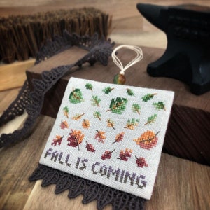 May include: A square cross-stitch ornament on light gray fabric featuring a pattern of small green, orange, and red autumn leaves. The text FALL IS COMING is stitched in dark purple at the bottom. It includes a thin hanging loop and dark lace trim.