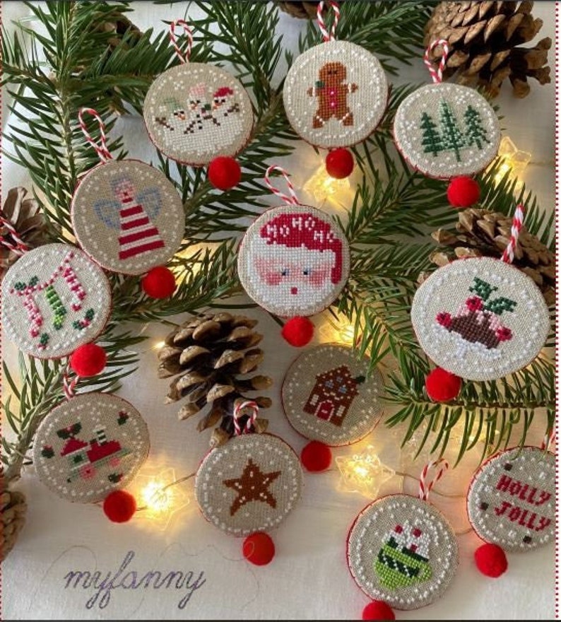 May include: A collection of round, embroidered Christmas ornaments with various festive designs, including a gingerbread man, Santa Claus, and a Christmas tree. Each ornament is adorned with a red pom-pom and a striped hanging loop, set against a backdrop of pine needles and pine cones.