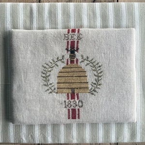 May include: A folded, off-white linen textile with a cross-stitched bee and beehive design. The design includes the word "BEE" and the year "1830". The textile is set on a striped, light green and white fabric.