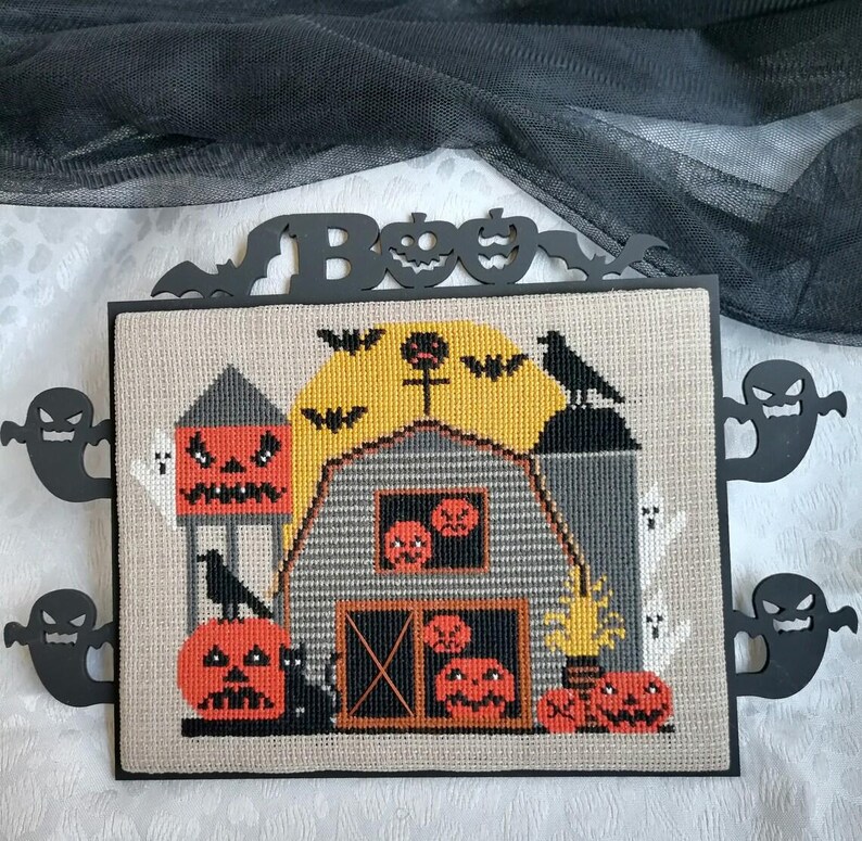 Może przedstawiać: Oprawiony haft krzyżykowy na Halloween przedstawiający scenę ze stodołą z dyniami, nietoperzami i wroną. Słowo "BOO" jest wycięte na g&oacute;rze, z dekoracyjnymi wycięciami duch&oacute;w po bokach. Stodoła jest szara z pomarańczowymi akcentami dyni.
