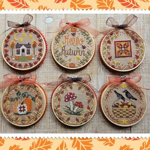 May include: Six embroidered hoop art pieces with autumn themes. Each features a different design, including a house, the text "Hello Autumn", a geometric pattern, pumpkins, mushrooms, and a basket with birds. They are framed in wooden hoops with red trim and tied with ribbons.