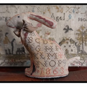May include: A stuffed fabric rabbit figurine featuring cross-stitch embroidery with the alphabet letters A through P and the year 1863 in muted red and brown tones. The rabbit has a small brown satin ribbon tied around its neck and a frayed tail.