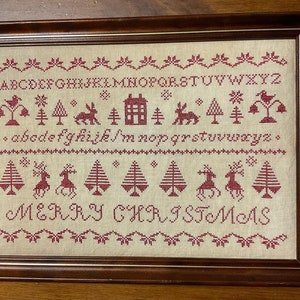 May include: A framed cross-stitch sampler in red and white. The sampler features a traditional alphabet, a row of winter-themed motifs, and the words "Merry Christmas".