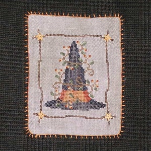 May include: A cross-stitch design of a witch's hat with a brown brim, black crown, and orange and green accents. The hat is surrounded by a brown frame and stars.