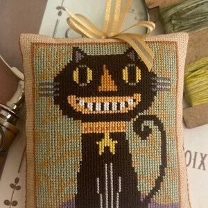 May include: Cross-stitched cat ornament with a smiling face, yellow eyes, and a star-shaped collar. The cat is black with a white-toothed grin, set against a green and tan background. A gold ribbon is attached for hanging.