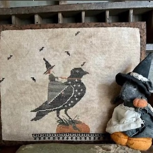 May include: A framed, stitched Halloween-themed artwork featuring a large black crow with white star accents, a witch, and bats. A plush crow wearing a witch hat and pumpkin body sits to the right. The artwork has a neutral colour palette with black, brown, and orange accents.