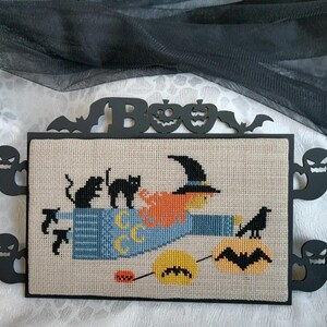 Może przedstawiać: Haft krzyżykowy z motywem Halloween przedstawiający wiedźmę na miotle, czarne koty i kruka. Grafika jest oprawiona w czarne wycięcia duch&oacute;w, nietoperzy i dyni. Nad grafiką znajduje się napis "BOO".
