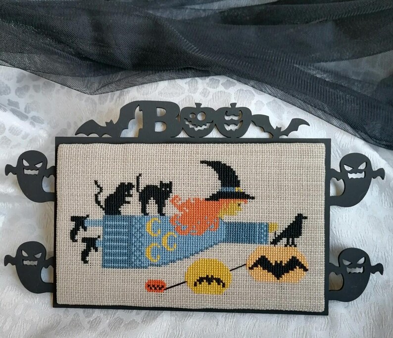 Może przedstawiać: Haft krzyżykowy z motywem Halloween przedstawiający wiedźmę na miotle, czarne koty i kruka. Grafika jest oprawiona w czarne wycięcia duch&oacute;w, nietoperzy i dyni. Nad grafiką znajduje się napis "BOO".