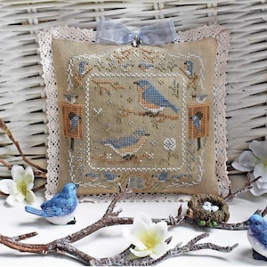 Puede incluir: Un cojín decorativo con un diseño de punto de cruz que representa pájaros azules posados en ramas. El cojín tiene un fondo beige, un ribete de encaje y una cinta azul claro. Figuras de pájaros azules y un nido con huevos están dispuestos alrededor.