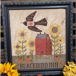 Op de afbeelding: Een ingelijst kruissteekkunstwerk met een bruine vogel in vlucht boven een rood gebouw met een damborddak. Gele zonnebloemen en pompoenen staan op de voorgrond. De tekst "BLACKBIRD INN" staat onderaan.