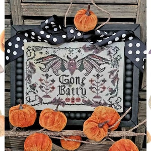 May include: A framed cross-stitch piece with the words "Gone Batty" and a bat design. The frame is black with a black and white polka dot ribbon. Orange velvet pumpkins and branches surround the frame, creating a Halloween theme.