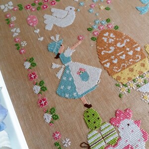 May include: A cross-stitch embroidery design featuring a girl in a blue dress holding a basket, a white dove, a large orange egg with a floral design, and two chicks with striped eggs. The background is a beige fabric with a floral pattern.