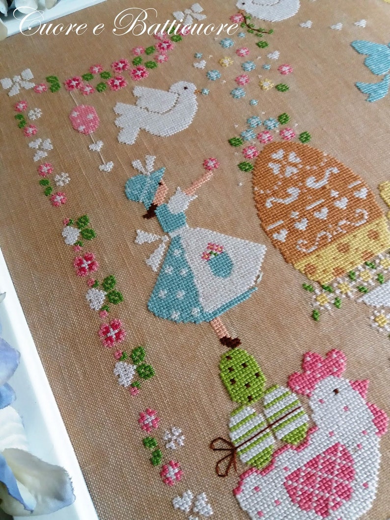 May include: A cross-stitch embroidery design featuring a girl in a blue dress holding a basket, a white dove, a large orange egg with a floral design, and two chicks with striped eggs. The background is a beige fabric with a floral pattern.