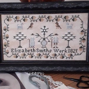 以下が含まれることがあります： 白い背景に黒い糸で刺繍された、額装されたクロスステッチのサンプル。サンプルには、木が2本、人物が2人、鳥が2羽、犬が2匹描かれています。テキスト「Elizabeth Smiths Work 1821」は黒い糸で刺繍されています。